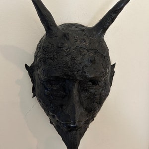 May include: A black, textured, devil mask with horns and a beard. The mask is made of a rough material and has a detailed face.