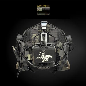 May include: A black tactical helmet with a camouflage pattern and a white patch featuring a hand symbol. The helmet has various attachments, including a headset and a communication device. The text "MSPEC" is visible at the top of the image.