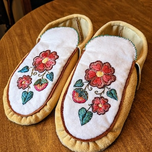 Op de afbeelding: Een paar gele suède mocassin-pantoffels met geborduurde bloemmotieven. De witte bovenkanten hebben rode en roze bloemen, groene bladeren en bruine stiksels. De pantoffels liggen op een houten oppervlak.