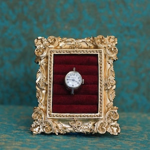 May include: A small silver-toned wristwatch with a white dial and black numerals is centered within a rectangular gold-colored frame featuring ornate scrollwork details. The watch rests against a deep red velvet insert with horizontal slots.