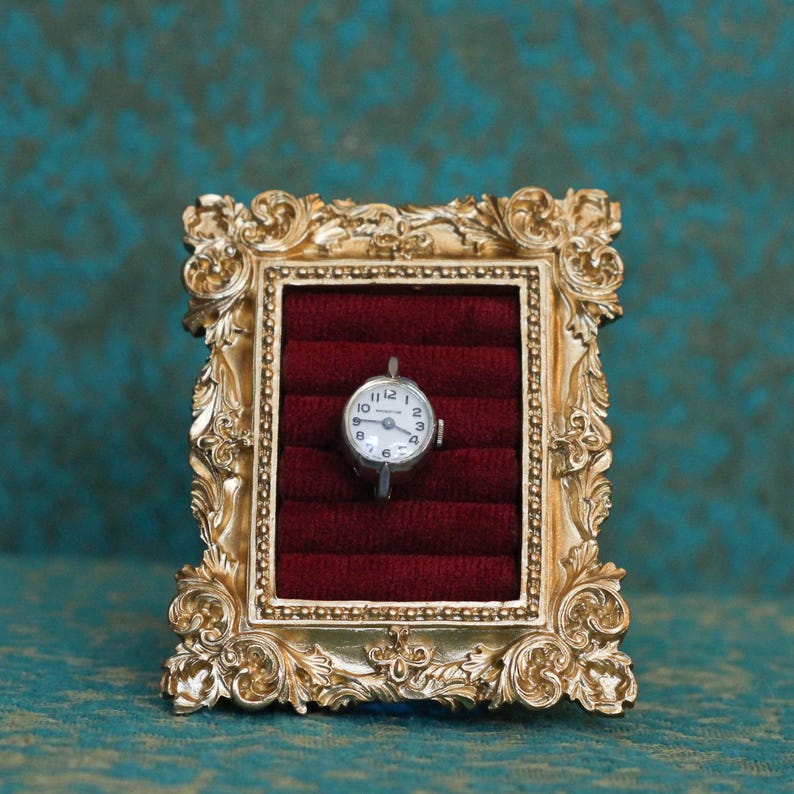May include: A small silver-toned wristwatch with a white dial and black numerals is centered within a rectangular gold-colored frame featuring ornate scrollwork details. The watch rests against a deep red velvet insert with horizontal slots.