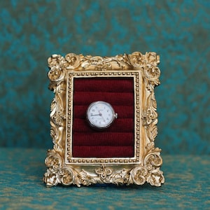 May include: A small, round, silver-toned analog watch with a white face and black numerals is centered within a rectangular, ornate gold-colored frame. The interior of the frame features deep red velvet padding with horizontal slots.