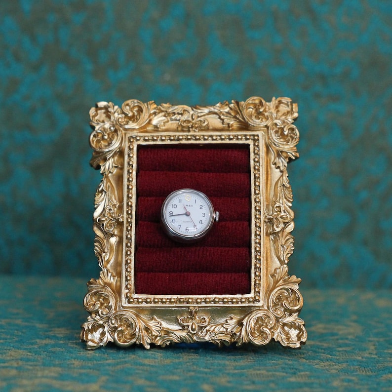 May include: A small, round, silver-toned analog watch with a white face and black numerals is centered within a rectangular, ornate gold-colored frame. The interior of the frame features deep red velvet padding with horizontal slots.