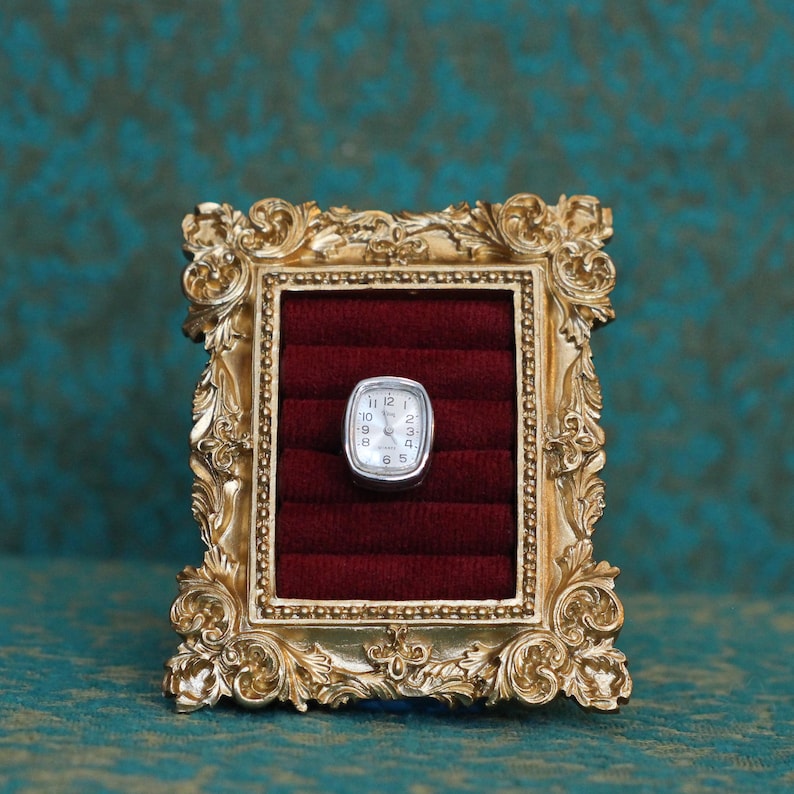 May include: A silver-toned wristwatch with a rectangular face and white dial featuring black numerals, displayed within a dark red velvet-lined ring holder set inside an ornate, gold-colored rectangular frame with intricate scrollwork details.