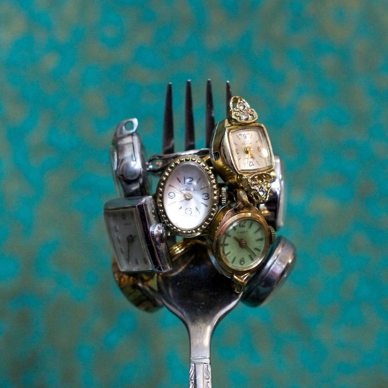 May include: A metal fork holds a cluster of various vintage wristwatches. The collection includes several small analog timepieces with gold and silver-toned metal cases, white and light green dials, and metallic hands, all balanced on the fork tines.
