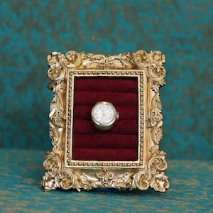 May include: A small, round, silver-toned wristwatch with a white face and dark hands is displayed inside a rectangular, ornate gold-colored frame. The interior of the frame is lined with deep red velvet, featuring horizontal slots for storage.