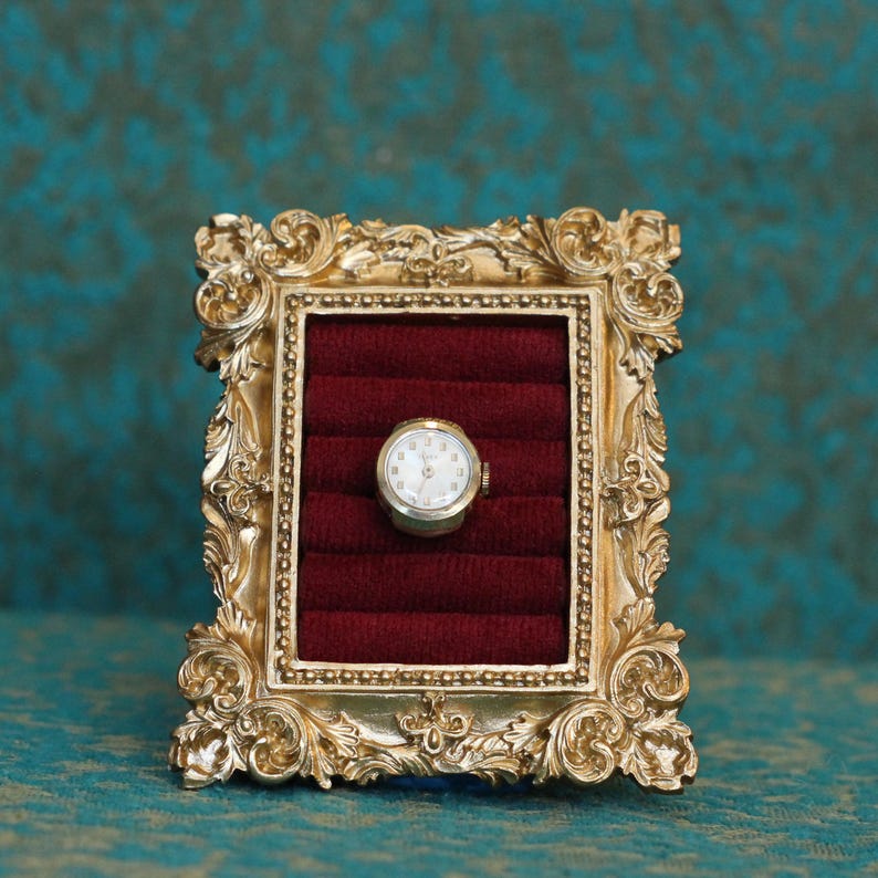 May include: A small, round, silver-toned wristwatch with a white face and dark hands is displayed inside a rectangular, ornate gold-colored frame. The interior of the frame is lined with deep red velvet, featuring horizontal slots for storage.