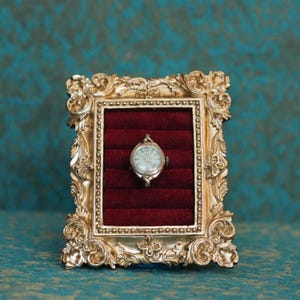 May include: A small silver-toned analog wristwatch with a round face and a winding crown, displayed inside a rectangular gold-colored frame featuring ornate scrollwork and a deep red velvet-lined interior with horizontal padded slots.