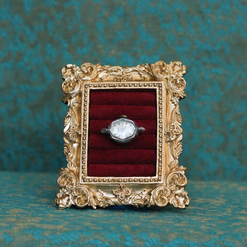 May include: A silver-toned wristwatch with a hexagonal face is displayed inside a small, ornate gold-colored frame. The watch rests against a deep red velvet-lined interior featuring horizontal slots, creating a structured display for the timepiece.