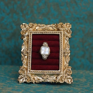 May include: A vintage-style gold-toned ring featuring a miniature watch face with a white dial and clear stones at the top and bottom, displayed within a rectangular gold ornate frame containing a deep red velvet ring insert for storage.