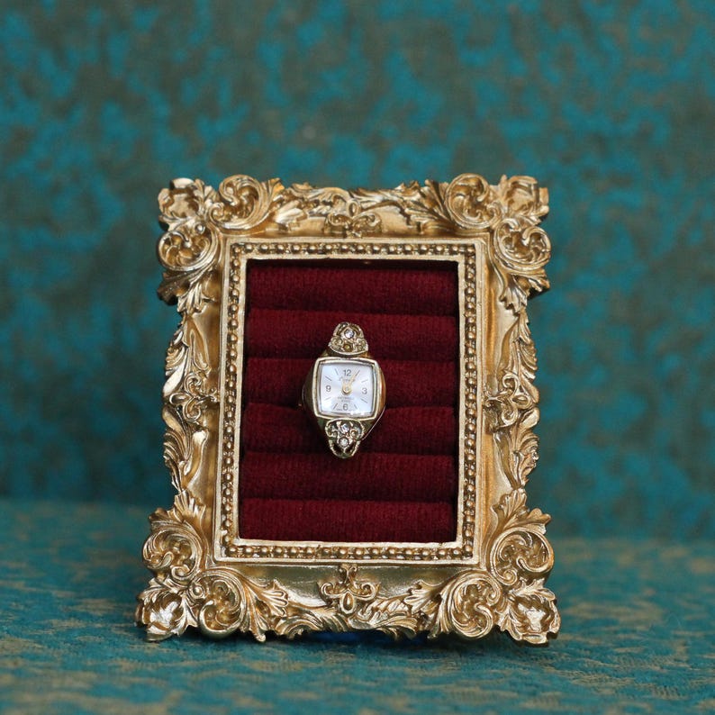 May include: A vintage-style gold-toned ring featuring a miniature watch face with a white dial and clear stones at the top and bottom, displayed within a rectangular gold ornate frame containing a deep red velvet ring insert for storage.