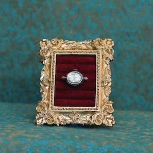 May include: A small, oval-shaped silver wristwatch with a white face and black numerals is displayed inside a rectangular, ornate gold-colored frame. The interior of the frame is lined with deep red velvet, featuring horizontal slots for storage.