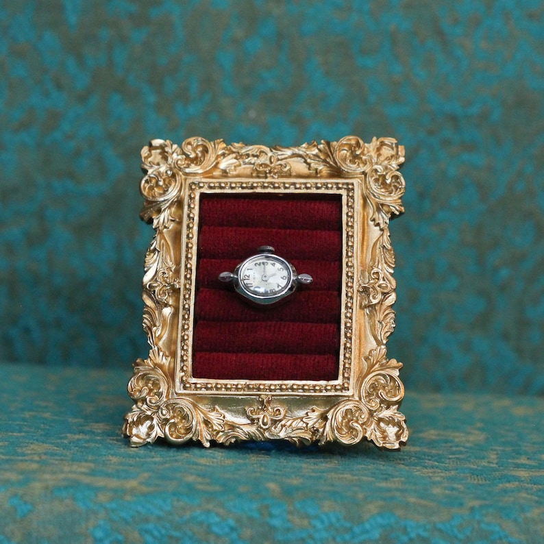 May include: A small, oval-shaped silver wristwatch with a white face and black numerals is displayed inside a rectangular, ornate gold-colored frame. The interior of the frame is lined with deep red velvet, featuring horizontal slots for storage.