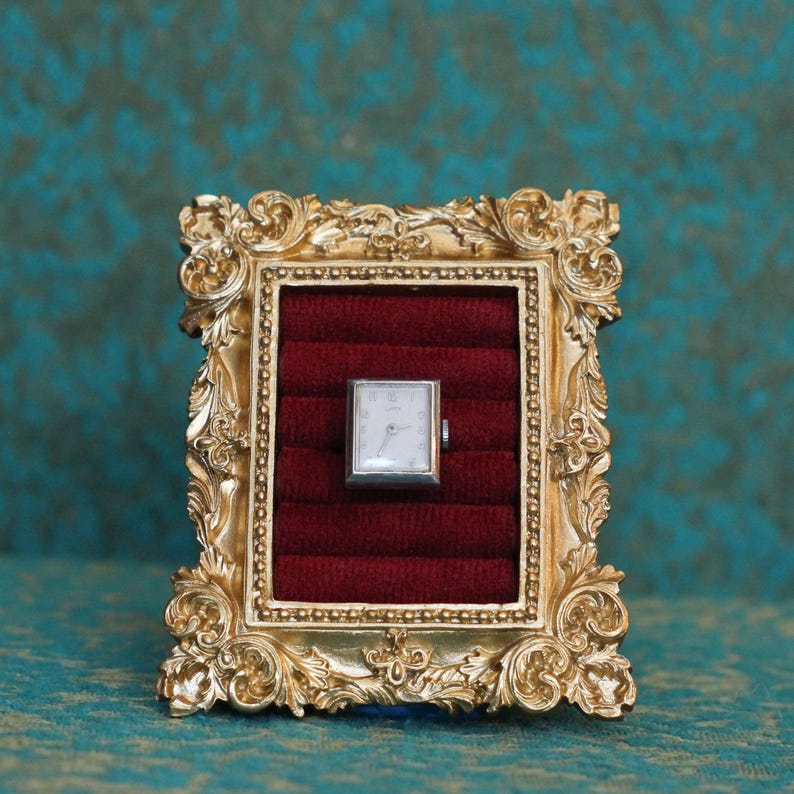 May include: A rectangular silver-toned wristwatch with a white face and black hands is displayed inside a gold-colored ornate frame. The watch is set against a deep red velvet-textured insert with horizontal ridges, creating a framed display.