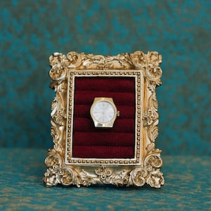 May include: A gold-toned ornate picture frame featuring a deep red velvet ring display insert. Centered within the velvet slots is a gold-colored wristwatch with a white circular face, thin gold hands, and a metallic gold-toned watch case.