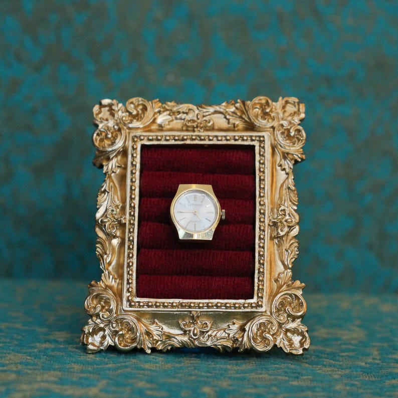 May include: A gold-toned ornate picture frame featuring a deep red velvet ring display insert. Centered within the velvet slots is a gold-colored wristwatch with a white circular face, thin gold hands, and a metallic gold-toned watch case.