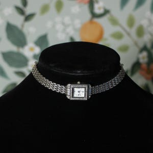 May include: A silver-toned watch choker necklace. The watch face is square with a white background and small black numbers. The watch face is surrounded by small, clear stones. The necklace is displayed on a black mannequin.