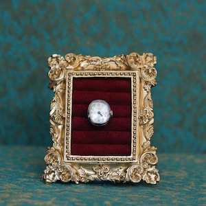 May include: A small, oval-shaped silver wristwatch with a white face and black numerals is centered within a rectangular, ornate gold-colored frame. The frame features intricate scrollwork and surrounds a deep red velvet ring display insert.