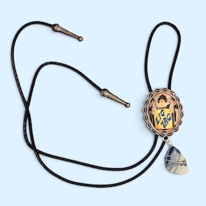 May include: A bolo tie featuring a black braided cord with silver-toned metal tips. The centerpiece is an oval frame with a scalloped edge containing an illustration of a figure holding a yellow sign with the blue text ZIG ZAG. A blue and white stone pendant hangs below.