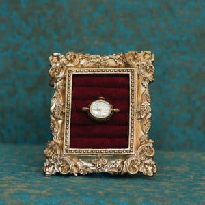 May include: A gold-toned ornate rectangular frame featuring intricate scrollwork and a beaded inner border. The center contains a deep red velvet insert with horizontal slots, holding a small gold-toned analog wristwatch with a white face.