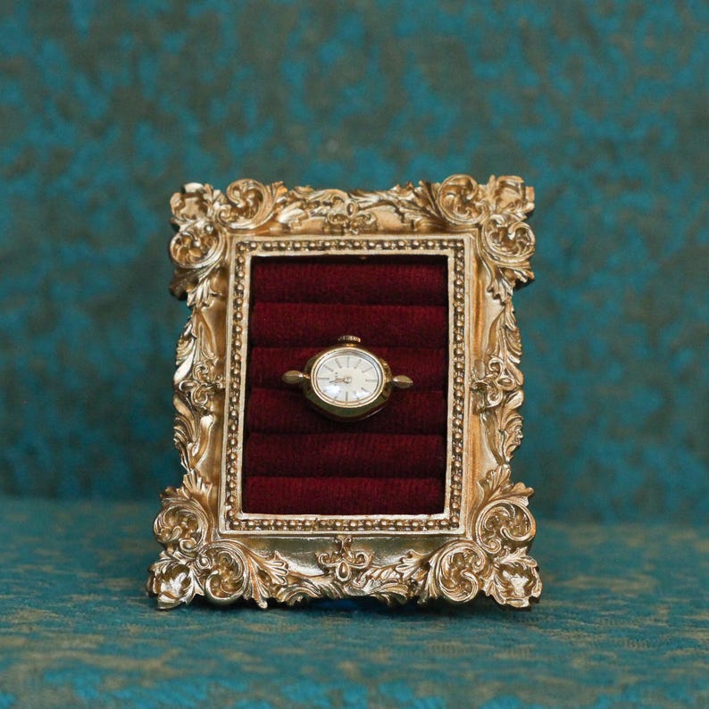 May include: A gold-toned ornate rectangular frame featuring intricate scrollwork and a beaded inner border. The center contains a deep red velvet insert with horizontal slots, holding a small gold-toned analog wristwatch with a white face.