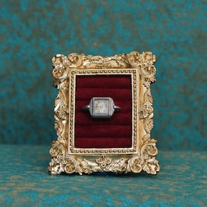 May include: A silver-toned square wristwatch head featuring a white dial with black numerals, displayed within a rectangular gold-colored ornate frame that contains a deep red velvet-textured insert with horizontal slots for holding jewelry.