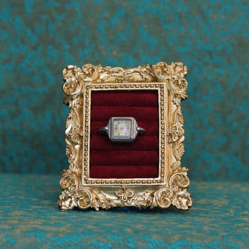 May include: A silver-toned square wristwatch head featuring a white dial with black numerals, displayed within a rectangular gold-colored ornate frame that contains a deep red velvet-textured insert with horizontal slots for holding jewelry.