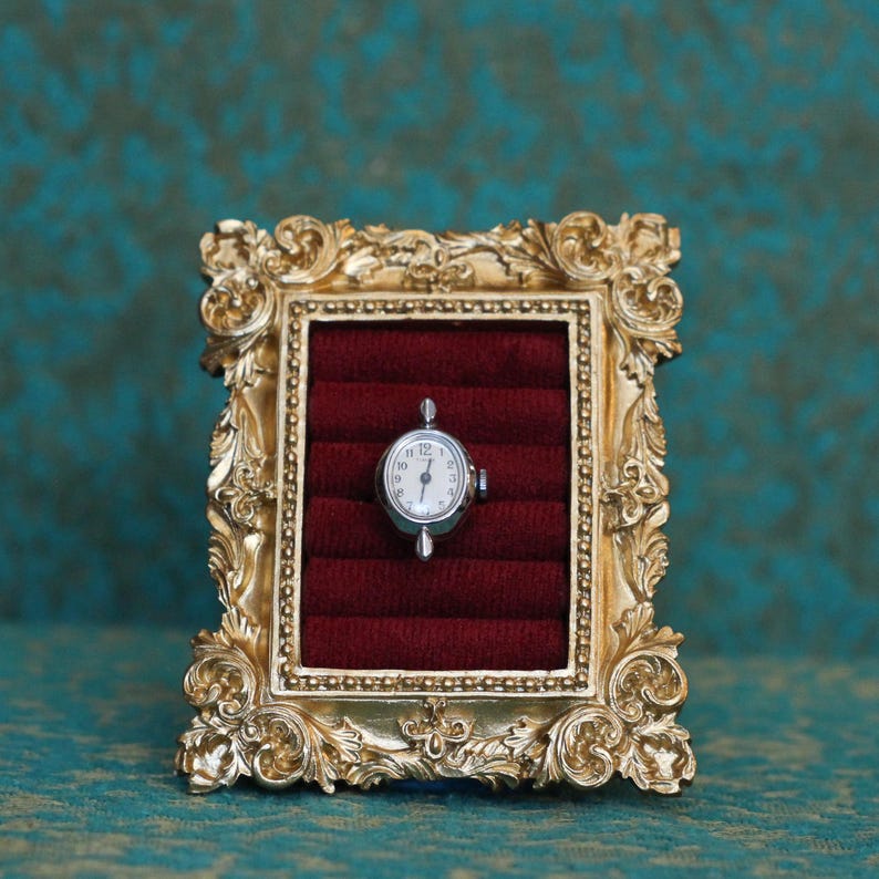May include: A small silver-toned oval watch face with a white dial and black numerals, displayed within a rectangular gold-colored ornate frame featuring a deep red velvet interior with horizontal slots for secure placement.