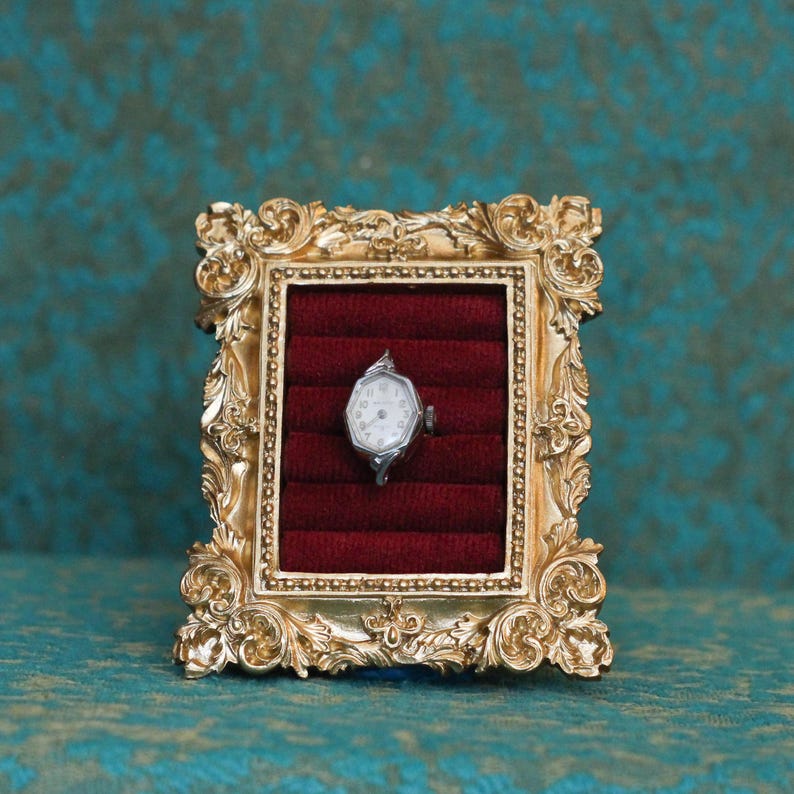 May include: A silver-toned wristwatch with a hexagonal face and white dial is displayed inside a rectangular, ornate gold-colored frame. The watch rests against a deep red, velvet-textured insert featuring horizontal padded slots.