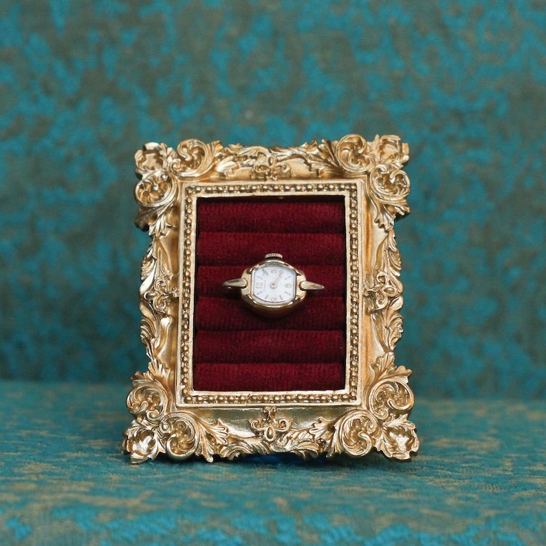 May include: A gold-toned ring featuring a small, white-faced analog watch with a metallic bezel. The ring is displayed within a rectangular, ornate gold frame that contains a deep red velvet insert with horizontal slots for jewelry storage.