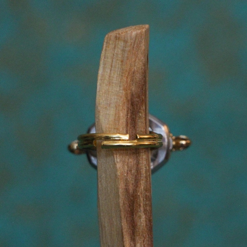 May include: A gold-toned adjustable ring band featuring a split design, positioned horizontally around a vertical piece of light brown wood with a visible grain texture. The ring has a metallic finish and a simple, open-ended circular structure.