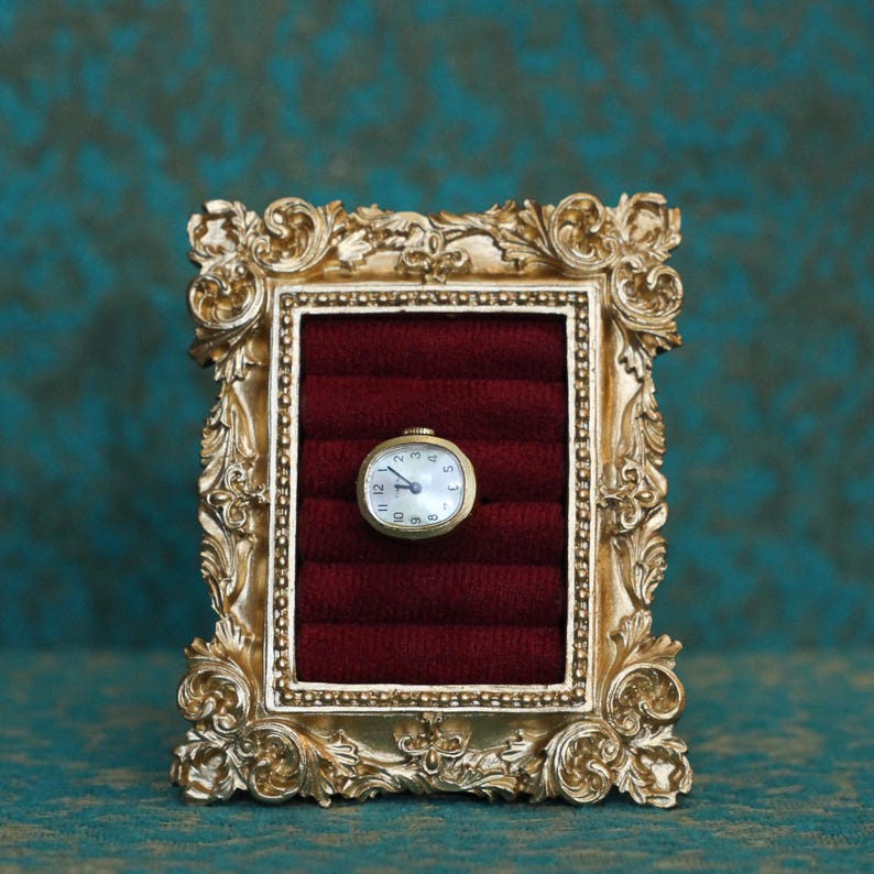 May include: A small gold-toned oval wristwatch with a white face and black numerals is centered on a deep red velvet ring display insert, which is housed within an ornate, rectangular gold-colored frame featuring intricate scrollwork details.