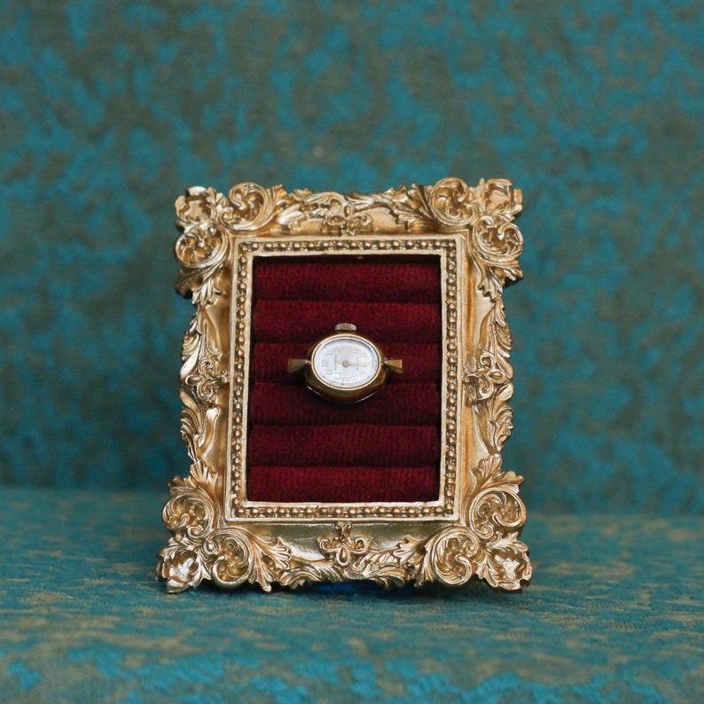 May include: A small gold-toned watch head with a white dial and black hands is centered within a rectangular, ornate gold-colored frame. The interior of the frame features deep red velvet padding with horizontal slots for secure display.
