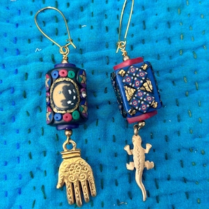 May include: A pair of handcrafted earrings with gold-colored ear wires. One earring features a dark blue cylindrical bead with a crescent moon design and a gold hand charm. The other has a gold lizard charm.