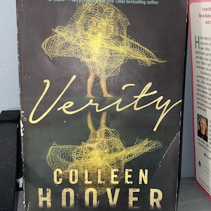 May include: A book cover for "Verity" by Colleen Hoover. The cover features a dark background with a yellow, abstract design and the title in gold lettering. The author's name and a quote are also visible.