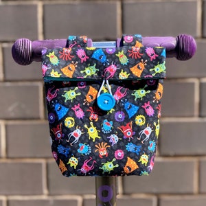 May include: A purple scooter handlebar with a black and white fabric bag attached. The bag has a colorful monster print and a blue button closure.