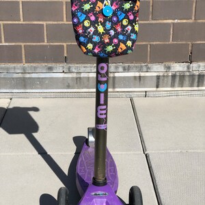 May include: Purple and black children's scooter with a monster-themed handlebar bag. The bag has a black background with colorful monsters printed on it. The scooter has a black handlebar and a purple deck.
