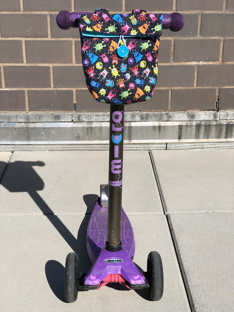 May include: Purple and black children's scooter with a monster-themed handlebar bag. The bag has a black background with colorful monsters printed on it. The scooter has a black handlebar and a purple deck.