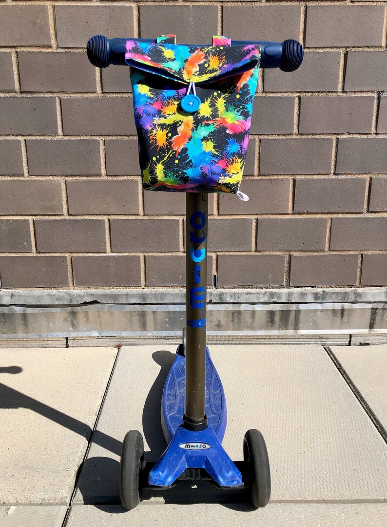 May include: A blue and black scooter with a colorful paint splatter design on the handlebar bag. The bag has a blue button and a white drawstring. The scooter has the word "micro" printed on the side.