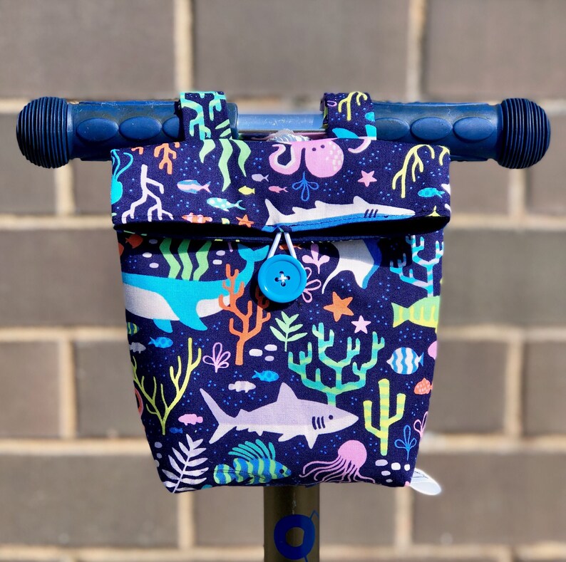 Peut inclure: Un guidon de scooter bleu avec un sac en tissu attach&eacute;. Le sac est d&eacute;cor&eacute; d'une sc&egrave;ne sous-marine color&eacute;e repr&eacute;sentant des requins, des coraux et des poissons. Le sac a une fermeture &agrave; bouton bleu.