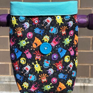 May include: A teal blue handlebar bag with a black fabric lining featuring a repeating pattern of colorful cartoon monsters. The bag has a blue button closure.