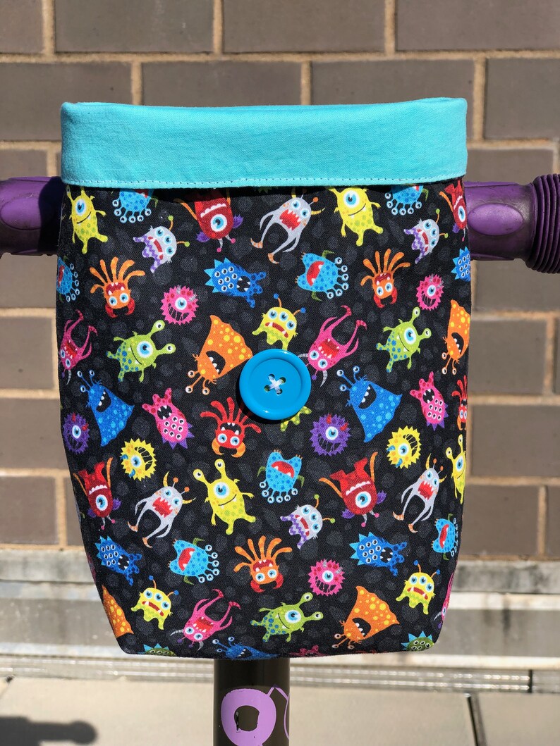 May include: A teal blue handlebar bag with a black fabric lining featuring a repeating pattern of colorful cartoon monsters. The bag has a blue button closure.