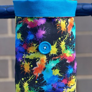 May include: A black fabric handlebar bag with a colorful paint splatter design. The bag has a blue button closure and a bright blue top.