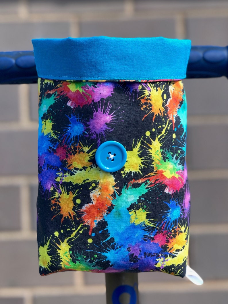 May include: A black fabric handlebar bag with a colorful paint splatter design. The bag has a blue button closure and a bright blue top.