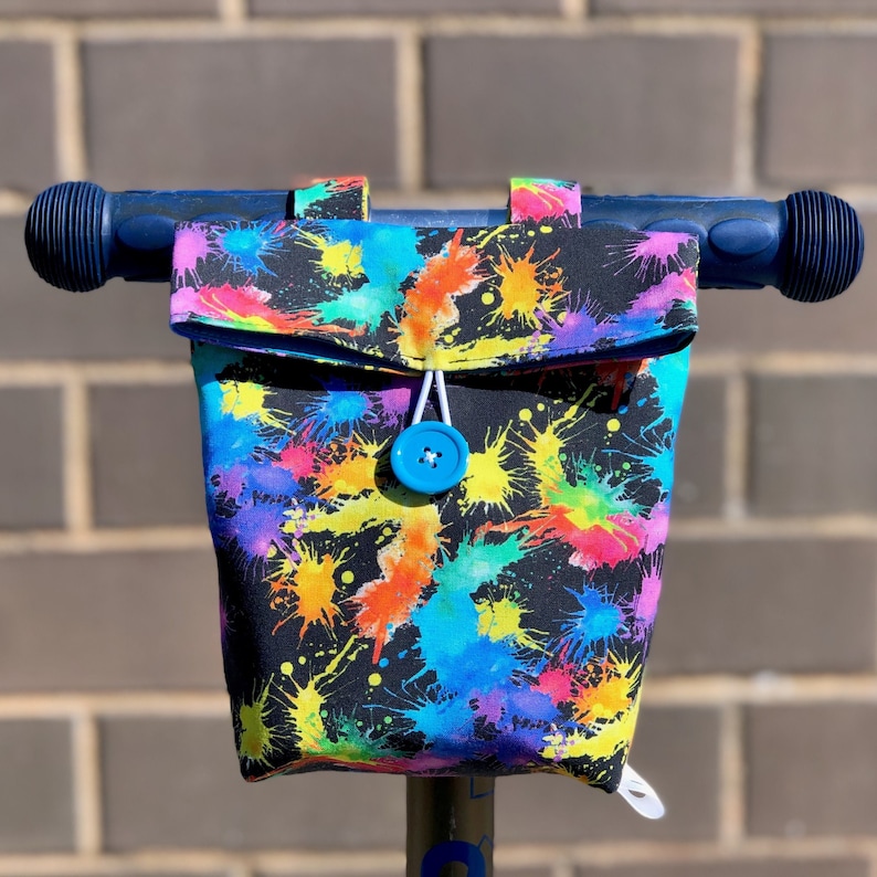 May include: A black handlebar bag with a colorful paint splatter design. The bag has a blue button closure and is attached to a scooter handlebar.