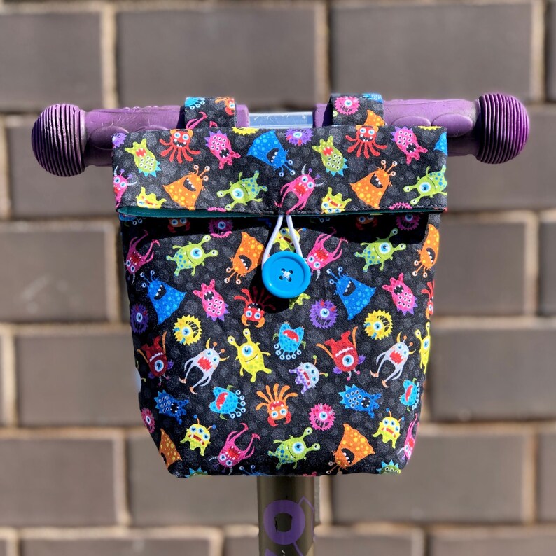 May include: A purple scooter handlebar with a black and white fabric bag attached. The bag has a colorful monster print and a blue button closure.