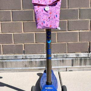 May include: A blue and black scooter with a pink fabric bag attached to the handlebars. The bag has a white zipper and a white button. The bag is covered in a pattern of science-related icons, including atoms, beakers, and test tubes.