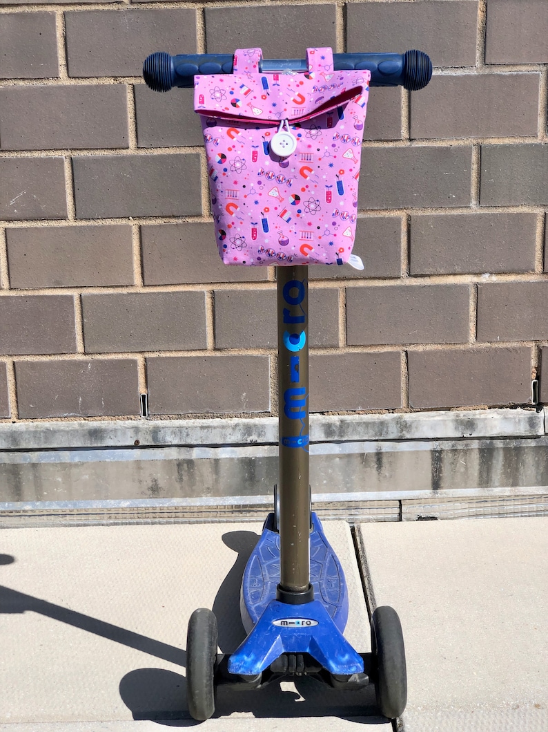 May include: A blue and black scooter with a pink fabric bag attached to the handlebars. The bag has a white zipper and a white button. The bag is covered in a pattern of science-related icons, including atoms, beakers, and test tubes.