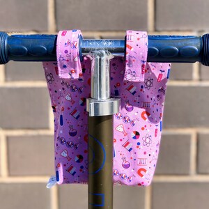 May include: Pink handlebar covers with a science theme, featuring beakers, atoms, and DNA strands. The covers are attached to a scooter handlebar.