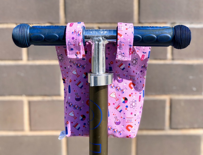 May include: Pink handlebar covers with a science theme, featuring beakers, atoms, and DNA strands. The covers are attached to a scooter handlebar.